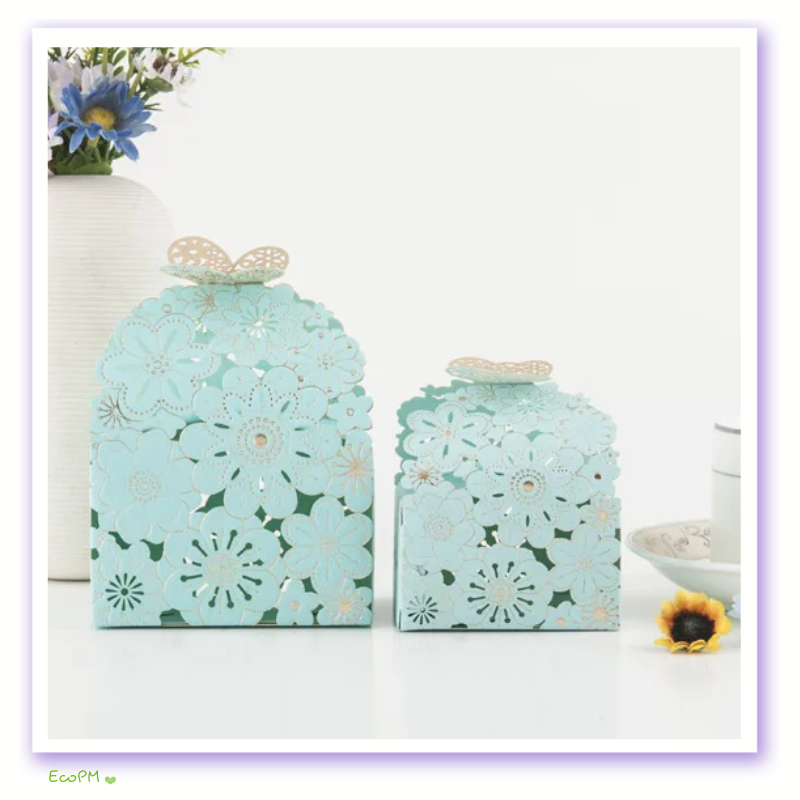 aqua-floral-laser-cut-favor-boxes.png Elegant aqua-colored favor boxes with intricate floral laser-cut design and butterfly closure displayed alongside a vase of blue flowers