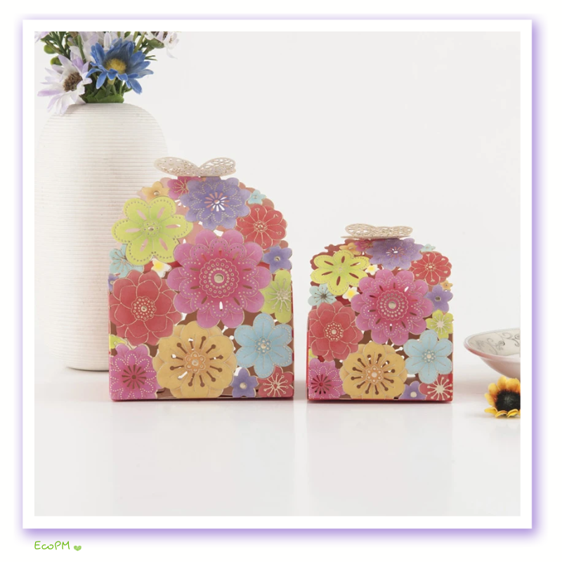 vibrant-floral-laser-cut-favor-boxes.png Two colorful favor boxes with intricate floral laser-cut patterns and butterfly closures, displayed alongside a white vase with a bouquet of wildflowers